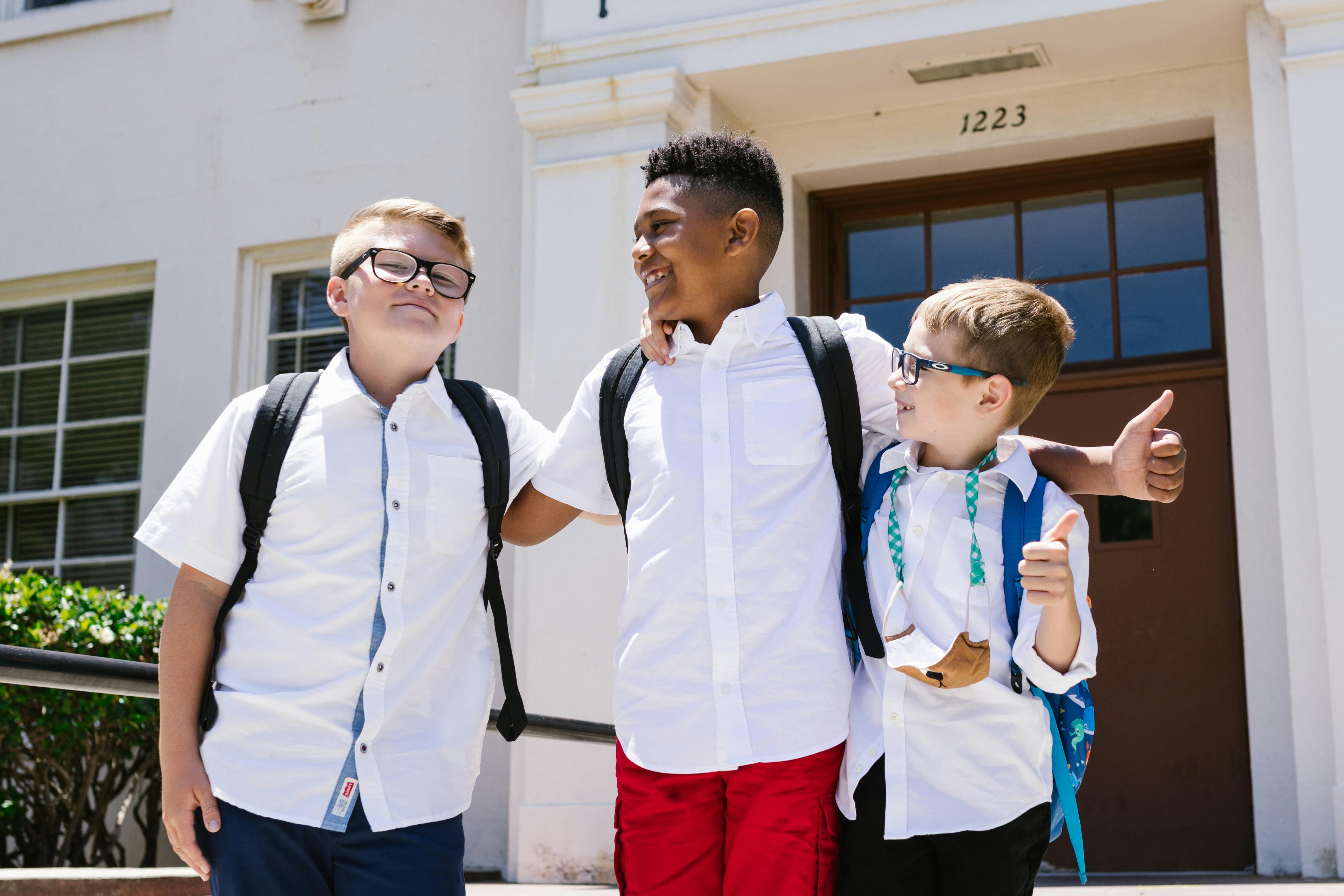 Three smiling schoolchildren with backpacks outside a school building, enjoying their first day back to school.